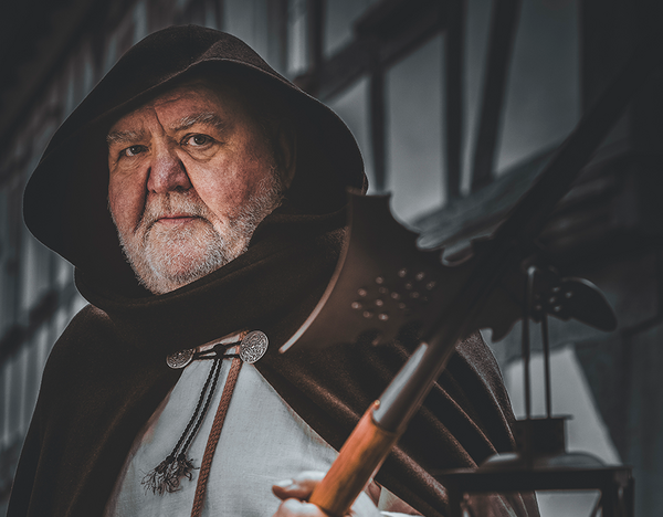 Das Bild zeigt eine Person in mittelalterlicher Kleidung, einschließlich eines Kapuzenumhangs und einem weißen Leinenhemd. Die Person hält in einer Hand eine Hellebarde und in der anderen eine Laterne mit einer brennenden Kerze. Der Stadtführer stellt einen Nachtwächter dar.