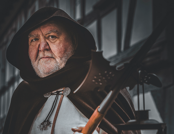 Das Bild zeigt eine Person in mittelalterlicher Kleidung, einschließlich eines Kapuzenumhangs und einem weißen Leinenhemd. Die Person hält in einer Hand eine Hellebarde und in der anderen eine Laterne mit einer brennenden Kerze. Der Stadtführer stellt einen Nachtwächter dar.