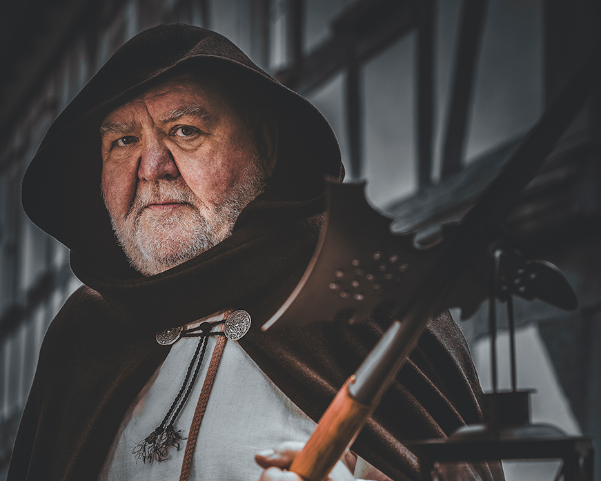 Das Bild zeigt eine Person in mittelalterlicher Kleidung, einschließlich eines Kapuzenumhangs und einem weißen Leinenhemd. Die Person hält in einer Hand eine Hellebarde und in der anderen eine Laterne mit einer brennenden Kerze. Der Stadtführer stellt einen Nachtwächter dar.