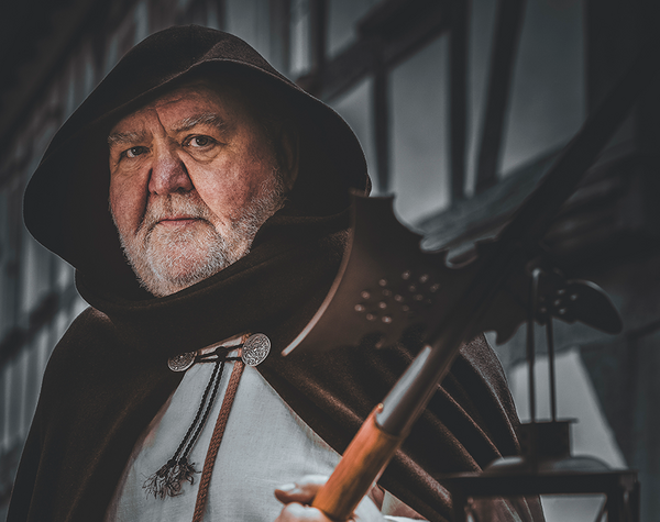 Das Bild zeigt eine Person in mittelalterlicher Kleidung, einschließlich eines Kapuzenumhangs und einem weißen Leinenhemd. Die Person hält in einer Hand eine Hellebarde und in der anderen eine Laterne mit einer brennenden Kerze. Der Stadtführer stellt einen Nachtwächter dar.